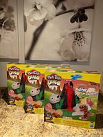 Three sealed Play-Doh Dino Crew boxed sets arranged on a counter under flower art.