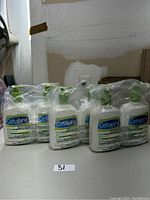 Six white Cetaphil lotion bottles with green pump tops packaged in plastic wrap, grouped in two sets of three bottles each on a table.