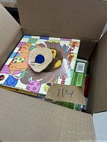Open cardboard box showing top book titled 'My First Book' with a beige paper tag marked 114 and attached plush lion hand puppet.