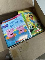 Top view of open box showing Peppa Pig's 'Peppa Goes Swimming' and 'Elly Socks' books on top, colorful children's books stacked below.