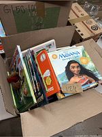 Photo of an open cardboard box filled with a variety of children's books stacked and slightly fanned out inside, showing the front covers of several titles including 'Moana'