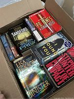 Top view of an open box with popular fiction best selling books packed inside, showing titles such as P.D. James A Certain Justice, John Sandford Silken Prey, Lisa Jackson Sinister, Lisa Jewell Found You, Stephen Hunter Point of Impact.