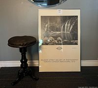 Art Deco piano stool with adjustable cast iron base and tufted dark leather seat beside framed black-and-white waterfall photography print leaning against wall.