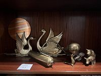 Five brass items displayed on a wooden shelf including two swan figurines, a cylindrical object with handle, a lamp with cord and spherical base, and a pig figurine.
