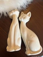 Photo of two white porcelain mid century modern cat figurines showing front and back views on a wooden surface with a white fur accent.