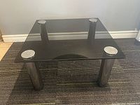 Square coffee table with smoked glass top and four chrome cylindrical legs, viewed from above on a carpeted floor.