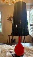Full view of the mid century modern table lamp showing the tall red base and large black tapered lampshade on a table with dining chairs in the background.