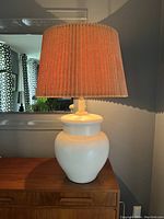 White glazed ceramic ginger jar lamp base with wide rounded shape, paired with a large pleated brown fabric shade. Positioned on wooden mid century style furniture against a dark wall with mirror reflecting the side view