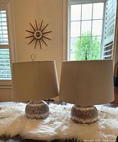 Pair of mid century modern ceramic table lamps shown with lampshades on displayed on a table with a furry white runner, sunburst clock in background.