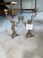 Pair of figural torchere candle holders shown on carpeted floor with furniture in background