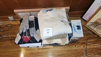 Folded knit sweaters including Tommy Hilfiger and Polo Ralph Lauren stacked on a wooden floor with a pair of gloves and a boxed electric clothing shaver.