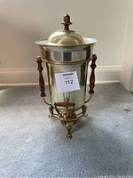 Front view of vintage coffee urn showing brass finish, wooden handles on sides, and brass spout with wooden lever under domed lid with wooden knob.