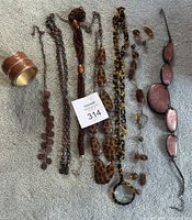 Full view of seven necklaces in brown tones and one wide cuff bracelet with brown leather-like material and metal edges arranged on carpet.