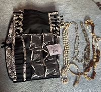 Photo showing five costume jewellery necklaces and an Ellen Tracey branded fabric jewellery hanging case laid flat on carpet. Necklaces include pearl-like beads, white and amber beads, and chain elements. Case has clear plastic pockets and multiple ties for organization.