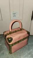 Front and side angled view of pink leather mini trunk bag with brass trim, two pink handles, and central lock.