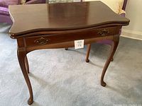 Wooden folding table with drawer and brass handle on carpeted floor with purple chairs in background.