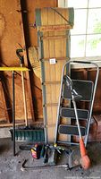 Photo showing vintage wooden Glacier snow sled, 3-tier step ladder, green snow shovel, orange electric weed wacker, chainsaw oil containers and crowbar arranged against a wall.