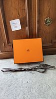 Orange Hermes box with lid and Hermes-branded brown ribbon on carpeted floor