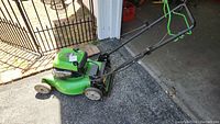Side view of green Lawnboy lawn mower showing engine and wheels, taken outside on pavement with a metal fence in background.