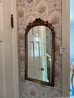 Full view of wooden framed arch mirror with carved decorative top, hanging on floral wallpapered wall.