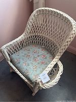 White wicker rocking chair with rounded back and woven lattice pattern, including blue floral seat cushion and side magazine basket.