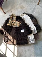 Overview of four women's coats laid out on a floor, showing different colors and textures including dark fur, embroidered cream coat, and checkered scarf.