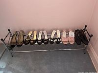 Six pairs of women's shoes placed on the top shelf of a two-shelf metal shoe rack against a pink wall and carpeted floor.