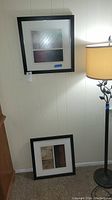 Two framed abstract wall art pieces shown, one hanging on wall and one resting on floor near lamp, both black framed and 19x19 inches.