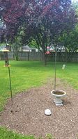 Two metal shepherd hooks and cement bird bath outdoors in garden bed with mulch, grass and trees in background.