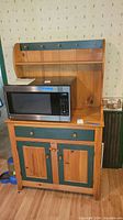 Front view of wood cabinet showing top shelf with hooks, microwave on top surface, drawer and double-door cabinet below.