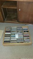 Wooden CD holder with many CD jewel cases arranged inside and in front on floor.