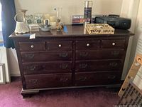 Front view of the 10 drawer dark wooden dresser with assorted items on top, showing brass and ceramic handles and some surface wear.