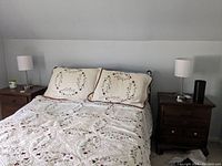 Photo of a bedroom with two identical table lamps on wooden bedside tables, both lamps have white cylindrical fabric shades and brushed metal bases.