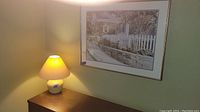 Photo of one table lamp lit on a wooden surface with one piece of framed wall art hanging above it, displaying an outdoor scene with a white picket fence and stone wall.