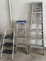 Three step ladders side by side, showing the 3-foot steel ladder, 4-foot aluminum ladder with blue top platform, and 6-foot aluminum ladder with metal top.