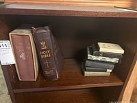 Shelf with one large Holy Bible with brown cover and gold embossed text, several smaller hymnals and Bibles stacked