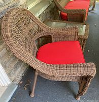 Side view of brown wicker style outdoor chair with red cushion seated on porch with stone wall behind