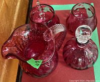 Four cranberry glass serving pieces on red cloth showing overall set with creamer, sugar, carafe with stopper
