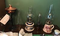 Four oil lamps displayed against a green wall, showing variety of pottery, glass, and metal bases with chimneys and accessories.