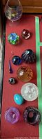 Top-down view of assorted glass pieces including paperweights and decorative glass objects arranged on a red surface, showing variety in size and color.