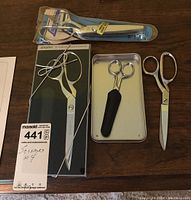 Full set of four scissors on wooden surface, showing all items including Gingher dressmaker shears in box, Singer scissors, small Gingher scissors with cover, Lamplough pinking shears