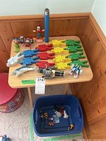 Full view of toys displayed on a small wooden table and blue bin on floor, showing wooden colored figures, dolphin, robots, frog, and small vehicles in bin