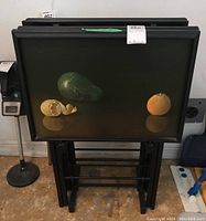 Front view of stacked black-framed TV trays with green fruit artwork showing three fruits on one tray: a green pear, peeled lemon, and orange.