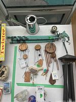 Overview of lot items on table including three wind chimes, three green metal garden stakes, metal watering can, bird hook, and some unrelated items in corner.