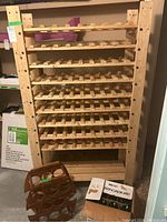 Full front view of large wood wine rack with multiple horizontal shelves. Smaller wooden rack and box of glass bottles visible at bottom front.