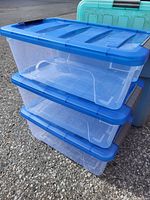 Three clear plastic storage containers stacked outdoors on pavement, each with a blue lid securely latched on top.