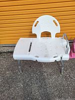 Photo shows a white plastic bath chair with contoured seat and backrest, grip handles on the backrest, and cutout in the seat for hygiene. The chair has adjustable aluminum legs with rubber feet for stability.