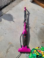 Photo of a pink Bissell stick vacuum cleaner laid on a concrete surface showing its full length and power cord wrapped around the handle.