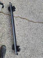 Pair of black roof car racks shown individually on pavement, featuring mounting clamps and ribbed surfaces.