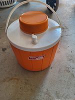 Photo of an orange plastic drink cooler jug with white lid, orange screw-on top cap, white spigot for drink dispensing, and a white carrying handle on a concrete floor.
